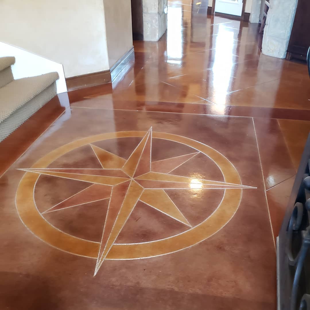 The image features a beautifully polished concrete floor with a decorative compass design scored into the surface. The compass motif, consisting of a star and circle, is etched into the concrete, providing an artistic touch. The floor has a high-gloss finish that reflects the surrounding space, enhancing the visual appeal of the design.