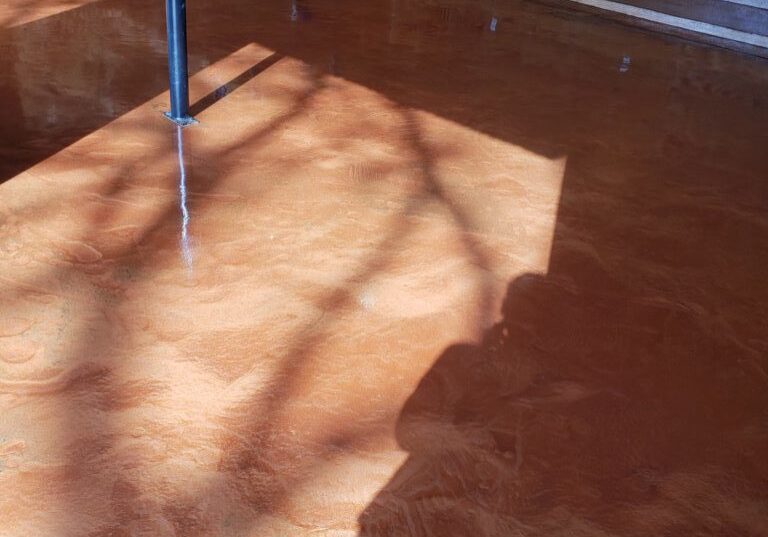 The image shows an interior space with a warm-toned, shiny epoxy floor. The reflective surface of the floor creates a mirror-like effect, casting reflections of objects and the sunlight streaming through a window. There's a distinct shadow of what appears to be a person taking the photo, contributing to the lived-in and personal feel of the space. The walls are partially clad in plywood, suggesting that this area may be under construction or renovation. Metal poles and plumbing fixtures are also visible, indicating that the space may serve a practical, possibly industrial or commercial function.
