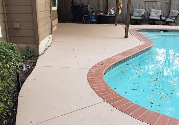 The image shows an outdoor residential patio area with a swimming pool. The patio is constructed with a beige-colored concrete surface that has a slip-resistant finish, suitable for poolside safety. A decorative brick border in a red tone neatly edges the curved outline of the pool, enhancing the aesthetic appeal of the backyard. The pool water appears clean but has some fallen leaves on the surface, suggesting a natural outdoor environment. Adjacent to the pool, there is a covered seating area with outdoor furniture, offering a comfortable space for relaxation or entertaining. The backyard is enclosed by a wooden fence, providing privacy for the residents.