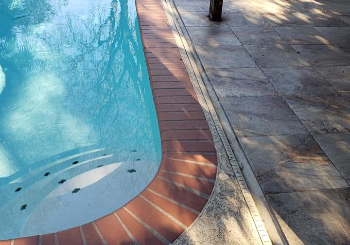 The image depicts a close-up view of a residential swimming pool area. The pool's clear blue water reflects the light and surroundings, creating a serene and inviting atmosphere. Surrounding the pool is a textured concrete deck, providing a safe, non-slip surface. A striking feature is the red brick coping that outlines the pool's edge, giving a classic and elegant contrast to the concrete. There is a single poolside post, possibly part of a fence or a support for a shade structure, casting a shadow on the deck. The presence of pool cleaning equipment in the background indicates regular maintenance, and the overall setting suggests a well-kept and enjoyable backyard leisure space.