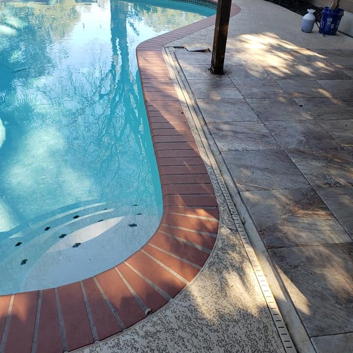 The image depicts a close-up view of a residential swimming pool area. The pool's clear blue water reflects the light and surroundings, creating a serene and inviting atmosphere. Surrounding the pool is a textured concrete deck, providing a safe, non-slip surface. A striking feature is the red brick coping that outlines the pool's edge, giving a classic and elegant contrast to the concrete. There is a single poolside post, possibly part of a fence or a support for a shade structure, casting a shadow on the deck. The presence of pool cleaning equipment in the background indicates regular maintenance, and the overall setting suggests a well-kept and enjoyable backyard leisure space.