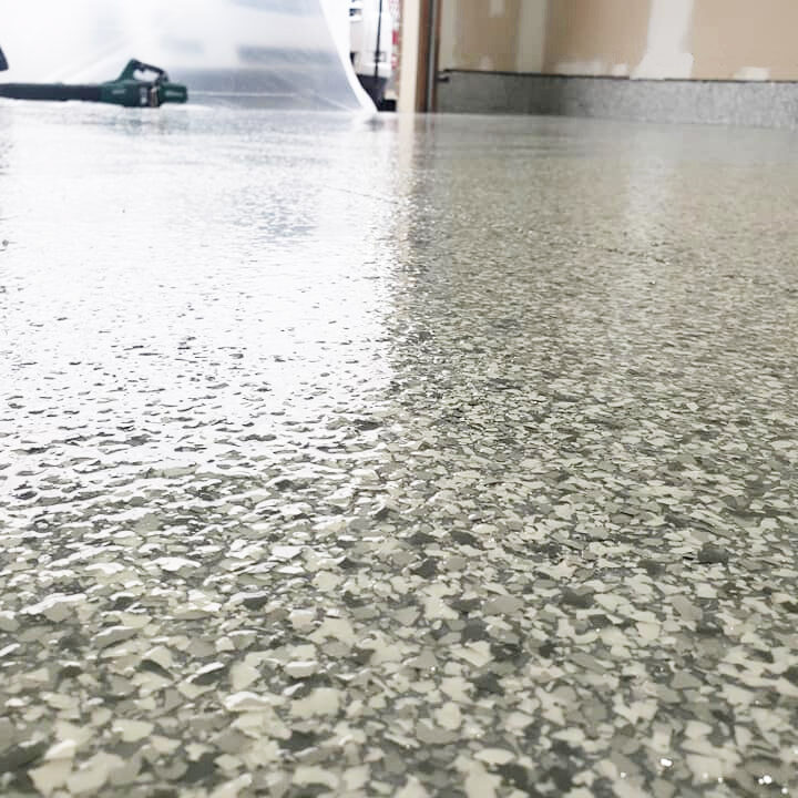 The image shows a close-up view of a glossy epoxy floor with a chip flake finish, creating a speckled effect with varying shades of gray and white chips scattered uniformly across the surface. The high-gloss finish of the epoxy coating reflects light and objects in the room, indicating a smooth and durable surface. In the background, the rest of the space is slightly blurred, with the outlines of furniture and possibly some construction equipment visible, suggesting that the area may be undergoing renovation or is used as a workspace. The floor's resilient and easy-to-clean nature, along with its aesthetic appeal, makes it a practical choice for both commercial and residential settings.